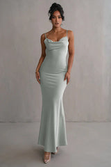 woman wearing a light green satin floor-length mermaid dress with spaghetti straps and a cowl neckline, standing against a grey wall