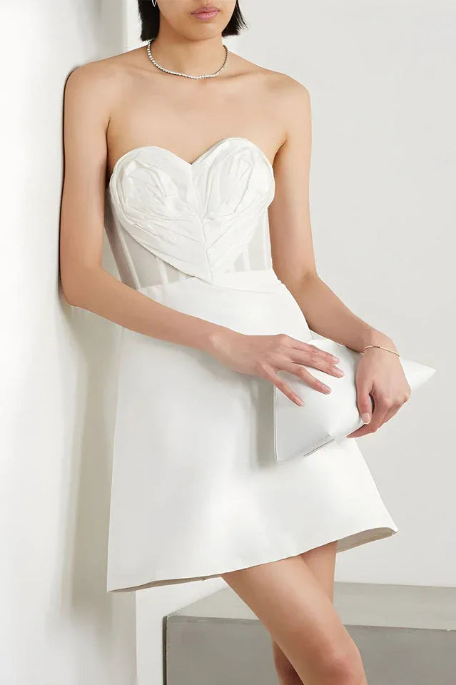 Woman wearing a white strapless mini dress with a heart-shaped applique on the bust, leaning against a wall and holding a clutch.