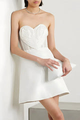Woman wearing a white strapless mini dress with a heart-shaped applique on the bust, leaning against a wall and holding a clutch.