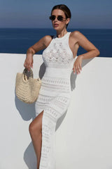 Woman leaning against a white wall wearing a white crochet knit maxi dress and sunglasses, holding a straw handbag.
