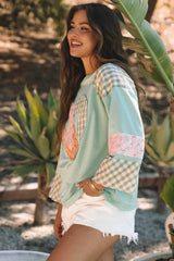Side view of a woman wearing a mint green long-sleeved top with a heart applique and plaid patchwork sleeves, smiling.