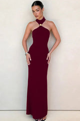 Woman wearing a burgundy sleeveless halter maxi dress with a gold-tone hardware detail at the neckline, standing against a neutral wall.