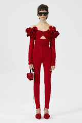 Full body shot of a model in a red off-the-shoulder top with ruffled floral shoulders and cutout, paired with red trousers, black sunglasses, crystal choker, and a red embellished handbag.