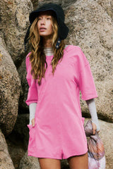 Close-up of a woman wearing a bright pink oversized tunic with short sleeves and pockets, layered with a grey long-sleeve top and a dark hat, outdoors.