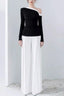 Model wearing a black long-sleeved top with an asymmetrical neckline and white wide-leg pleated pants.