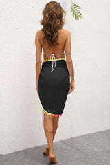 Rear view of a woman wearing a white bikini top and a black sarong with colorful pom-pom trim.