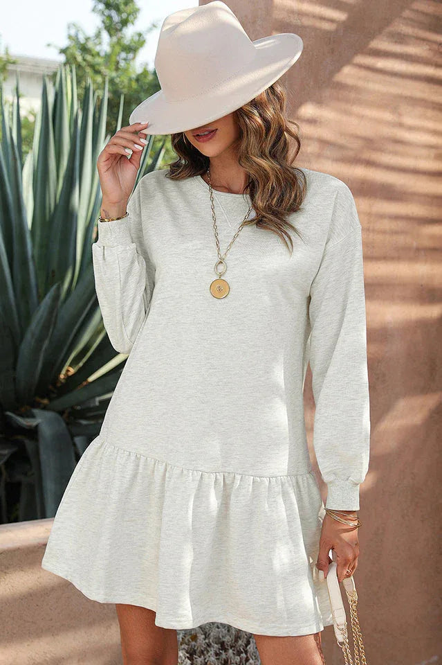 Full-body shot of a woman in a light grey long-sleeved drop-waist dress with a ruffled hem, wearing a cream fedora hat and white cowboy boots.