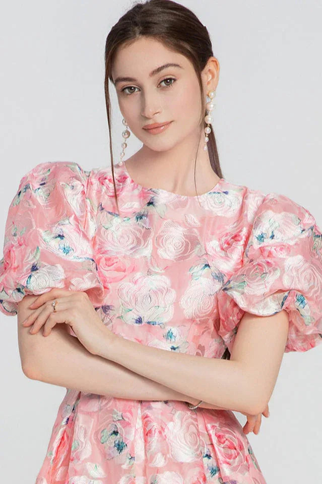 Close-up of a woman's upper body wearing a pink floral dress with puff sleeves and pearl earrings.