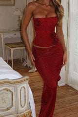 Side view of a woman in a red sparkly strapless corset top and maxi skirt, with a bed and vintage furniture in the background.