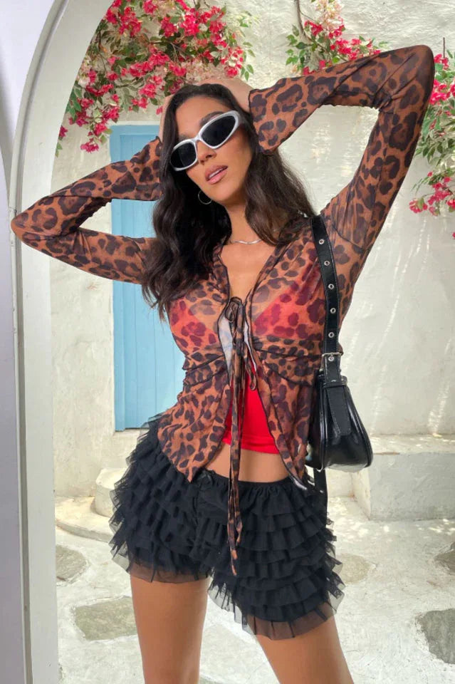 Woman posing with hands on head wearing a leopard print sheer mesh top and black ruffled shorts.