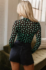 Rear view of a woman wearing a black long-sleeved mesh bodysuit with a green shamrock print and black denim shorts.