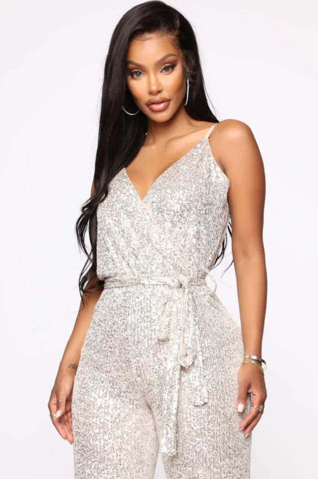 Close-up front view of a woman wearing a silver sequined sleeveless wrap jumpsuit with a self-tie waist belt.