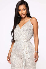 Close-up front view of a woman wearing a silver sequined sleeveless wrap jumpsuit with a self-tie waist belt.