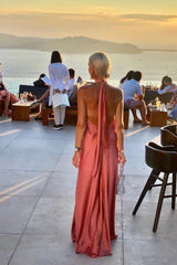 Back view of a woman in a coral satin halter maxi dress at an outdoor event during sunset.