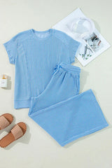 Flat lay of a blue ribbed short-sleeve top and wide-leg pants with sandals, headphones, and a perfume bottle.