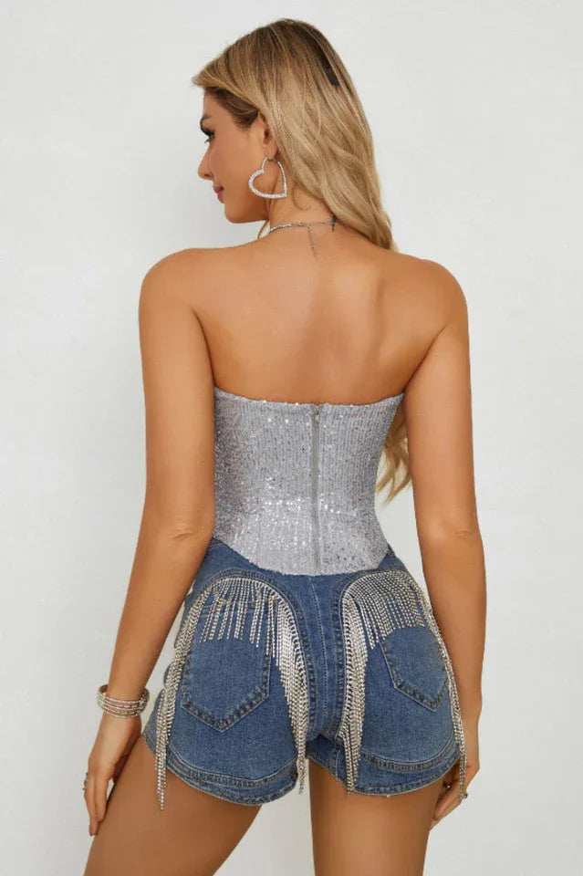 Full body shot of a woman in a silver sequined strapless corset top, blue denim shorts, and clear heels, holding a small silver handbag.