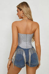 Full body shot of a woman in a silver sequined strapless corset top, blue denim shorts, and clear heels, holding a small silver handbag.