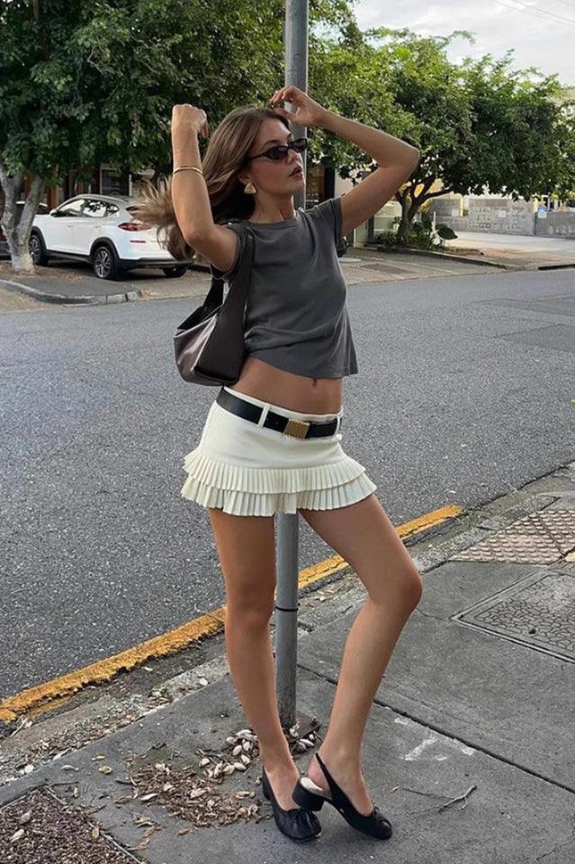 Model wearing a white pleated mini skirt with a grey t-shirt, black belt, and black slingback heels.