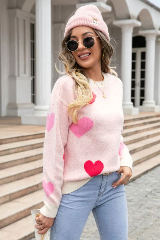 A woman wearing a pink heart print sweater and a pink beanie, wearing sunglasses and holding a purse.