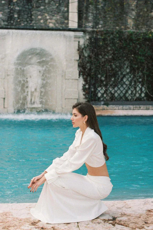 Woman in a white satin two-piece set crouching by a pool.