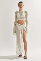 Model wearing a white sheer knit cropped long-sleeve top, bikini, and a matching white sheer knit maxi skirt with a high slit, showcasing the side.