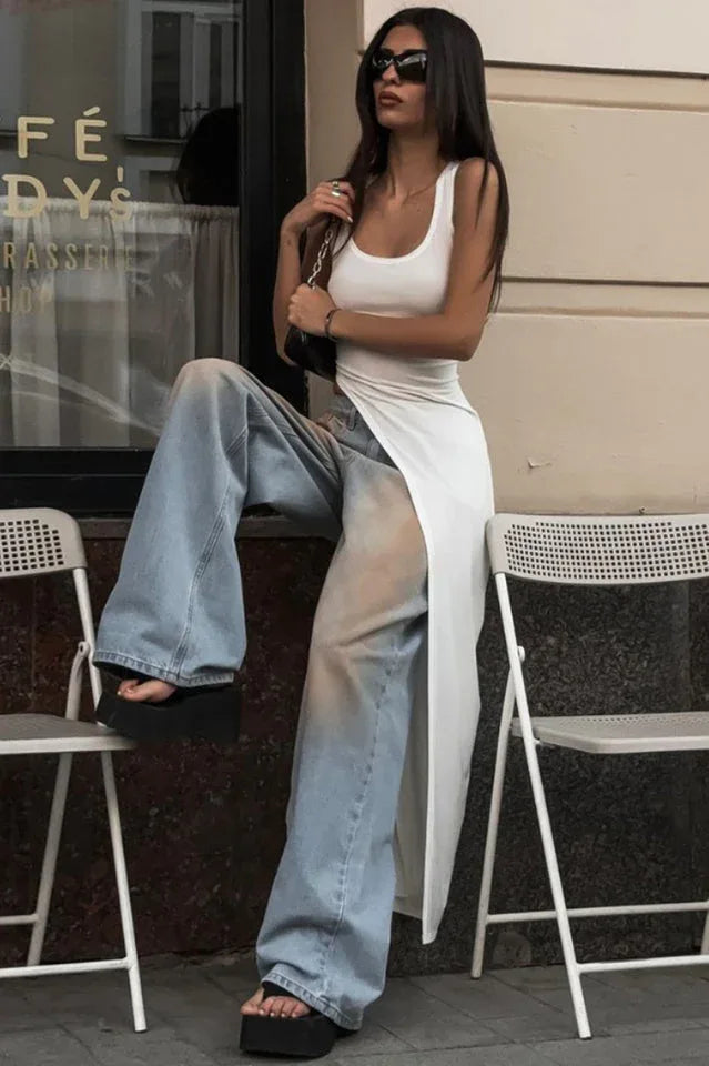 Woman sitting on a chair wearing a white sleeveless maxi dress with a high slit over light blue wide-leg jeans and black platform sandals