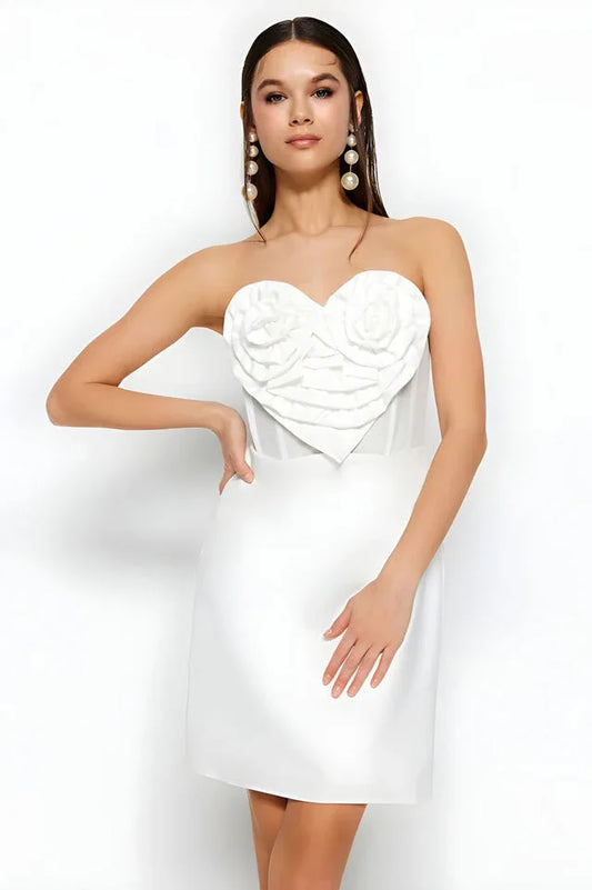 Woman wearing a white strapless mini dress with a heart-shaped applique on the bust, posing with her hand on her hip.