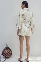 Woman wearing a white floral print button-down shirt with puffed sleeves and matching high-waisted shorts, back view, standing next to a decorative net ball