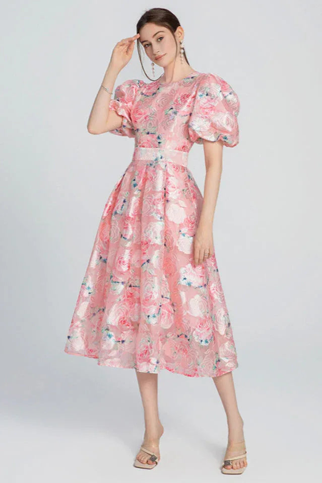Woman standing in a pink floral midi dress with puff sleeves and a belted waist, showing the full silhouette.