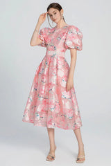 Woman standing in a pink floral midi dress with puff sleeves and a belted waist, showing the full silhouette.