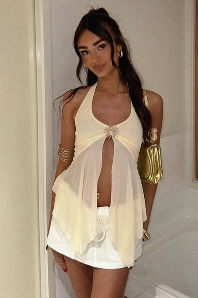 Woman wearing a cream halter top with a sheer, flowing overlay and a sunburst embellishment, accessorized with gold bracelets and a gold handbag.