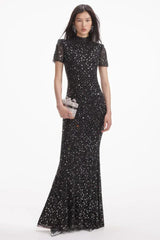 Model wearing a black sequined floor-length gown with a high neckline and short sleeves, with a silver clutch.
