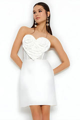 Woman wearing a white strapless mini dress with a heart-shaped applique on the bust, with her hands behind her back.
