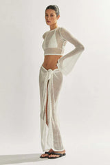Model wearing a white sheer knit cropped long-sleeve top, bikini, and a matching white sheer knit maxi skirt with a high slit, angled view.