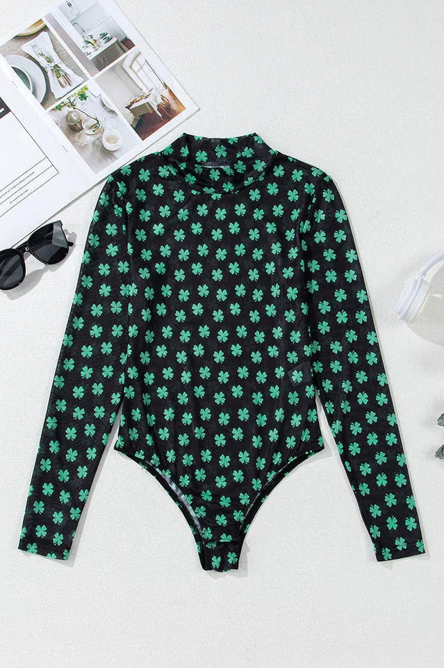 A black long-sleeved mesh bodysuit with a green shamrock print laid flat, with sunglasses and a magazine nearby.