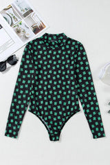 A black long-sleeved mesh bodysuit with a green shamrock print laid flat, with sunglasses and a magazine nearby.