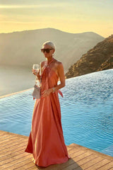 Woman in a coral satin halter maxi dress holding a drink by an infinity pool at sunset.