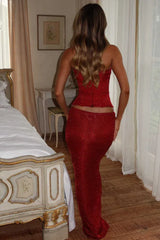 Back view of a woman in a red sparkly strapless corset top and maxi skirt, with a bed and vintage furniture in the background.