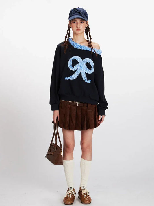 Model wearing a black off-the-shoulder sweatshirt with a large light blue appliqué bow on the front and ruffled blue trim.