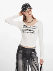 Front view of a woman wearing a white long-sleeved graphic t-shirt, grey distressed jeans with a black fringed belt, and a black beaded necklace, with her hands on her baseball cap.