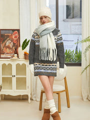 Woman wearing a gray knit sweater dress with a Fair Isle pattern, a multi-colored fringed scarf, a cream beanie, white gloves, and white leg warmers.