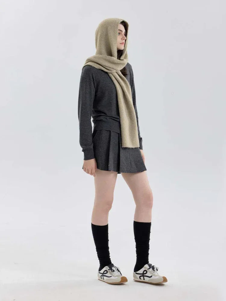 Full body shot of a model wearing a gray sweater and pleated skirt with a beige hooded scarf, black socks, and sneakers.