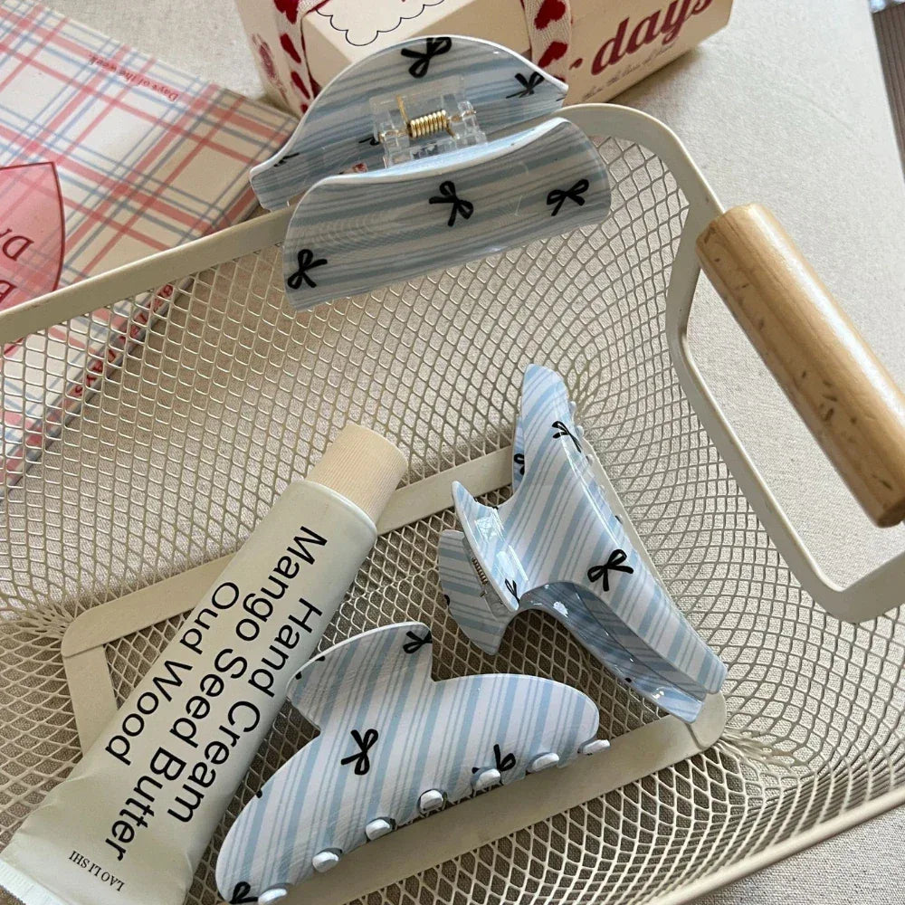 Three light blue and white striped hair claw clips with black bow prints inside a white mesh basket with a tube of hand cream and a mug.