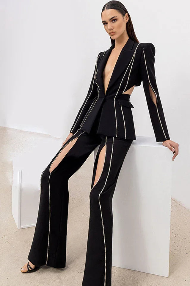 Woman wearing a black suit with cut-out details and decorative trim, posing on a white cube.