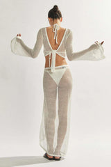 Back view of a model wearing a white sheer knit cropped long-sleeve top with a tie closure, bikini, and a matching white sheer knit maxi skirt.
