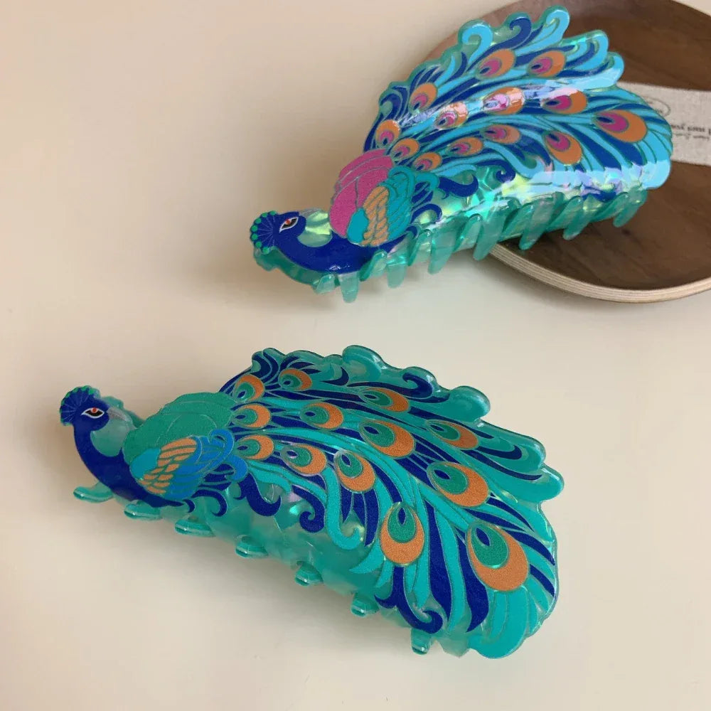 Two peacock hair claw clips, one in the foreground and one in the background, showcasing their iridescent blue and green colors and detailed feather patterns.