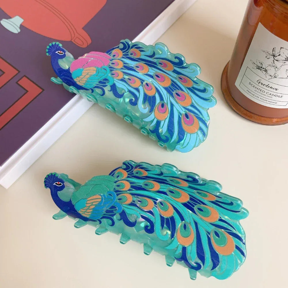 A close-up of two peacock hair claw clips, one with pink accents and the other with orange accents, placed near a scented candle and a book.