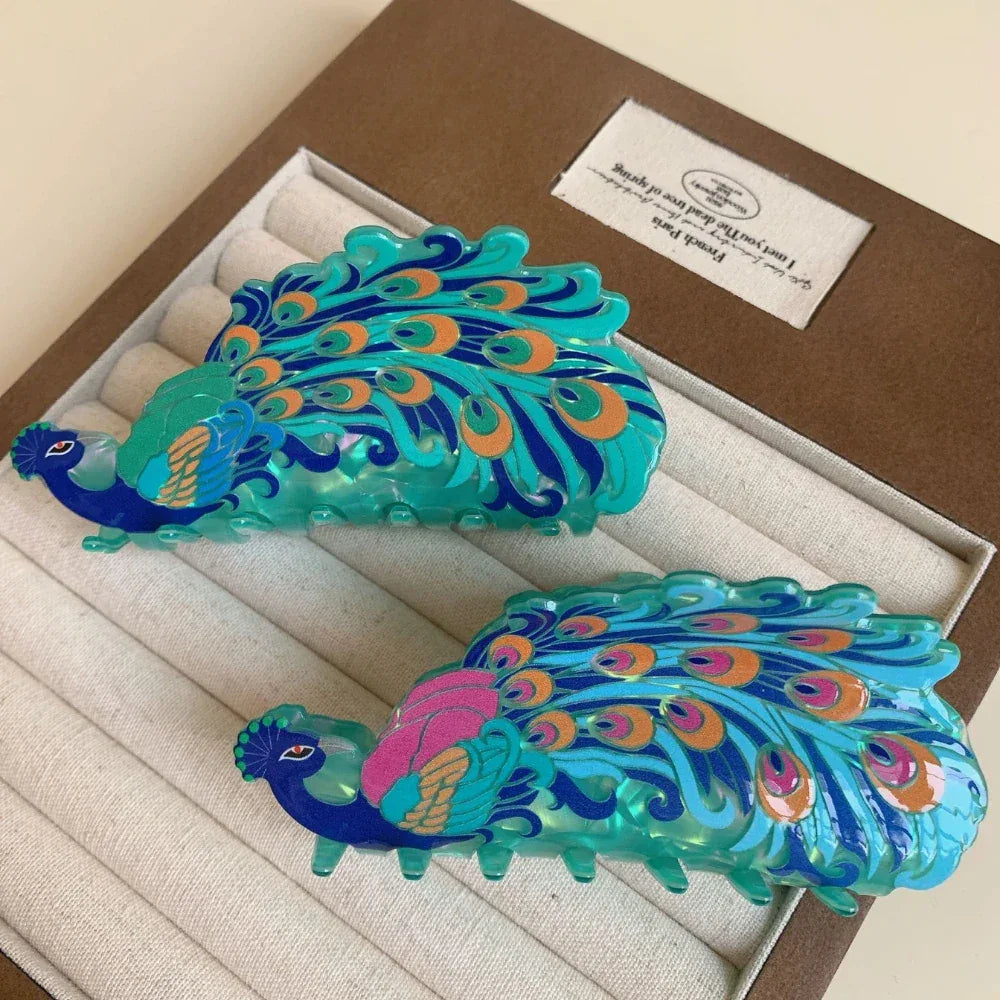 A pair of peacock hair claw clips, one with pink accents on the feathers and the other with orange accents, nestled in a jewelry box.
