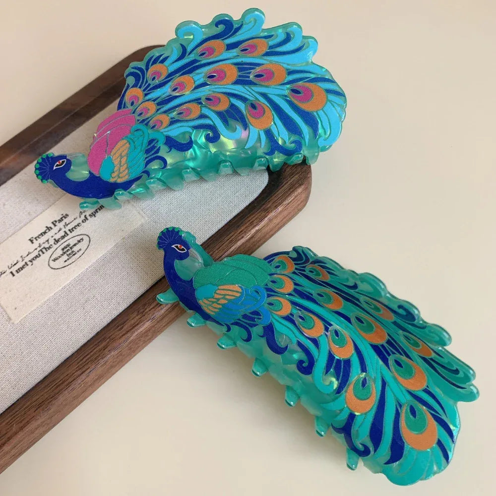 A pair of peacock hair claw clips arranged on a wooden tray, highlighting their detailed design and vibrant colors.