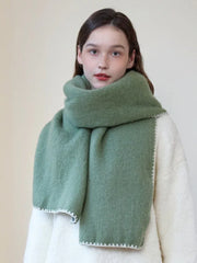 Woman wearing an oversized green scarf with a white fringed edge and a cream-colored sweater, looking forward.